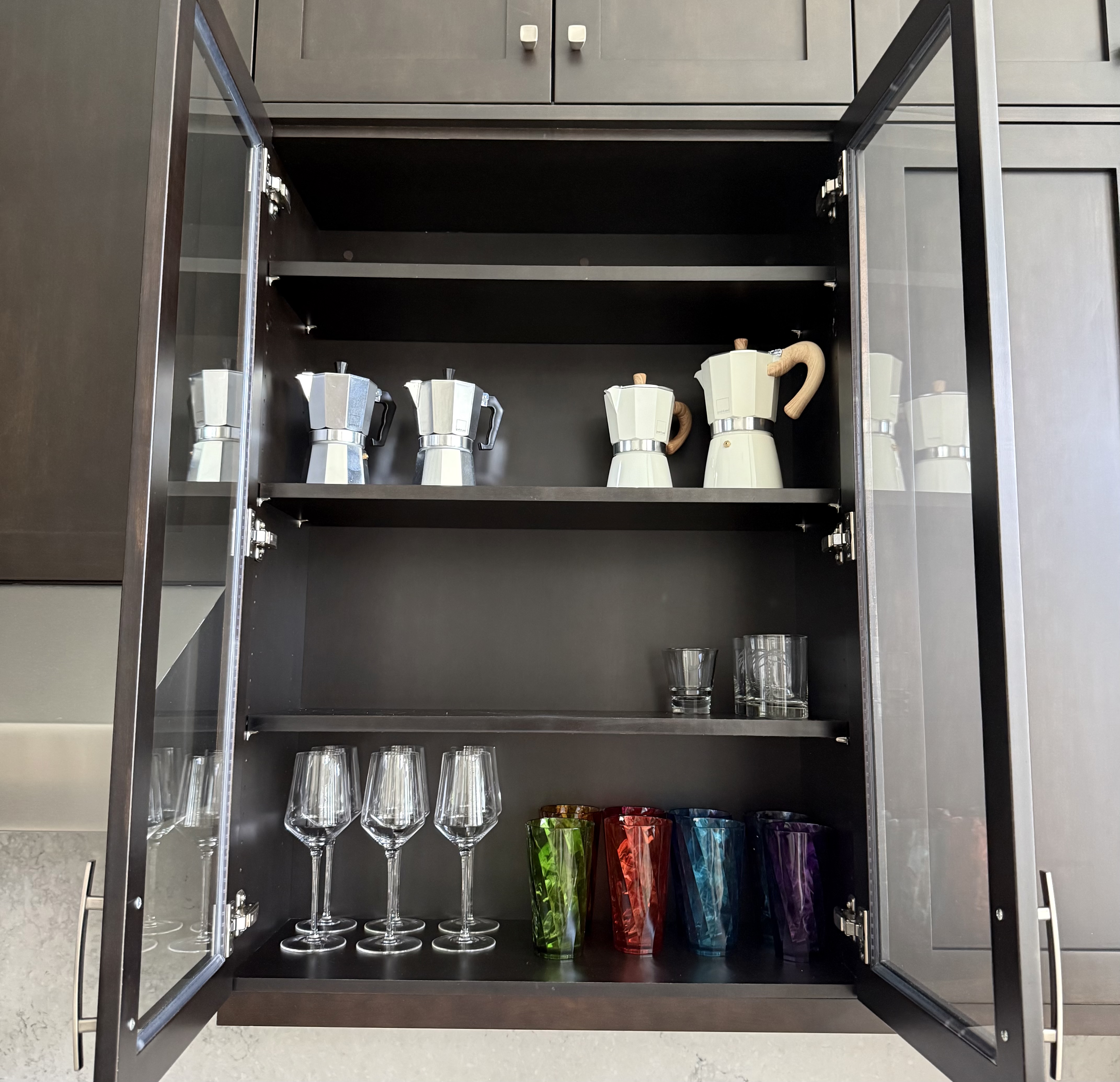 Glass-front bar cabinet organized with moka pots, crystal stemware, and colorful glassware